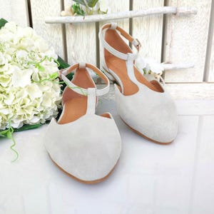 May include: A pair of light gray suede T-strap flats with ankle straps and brown soles. The shoes are displayed on a white surface with a white floral arrangement in the background. The flats have a classic, elegant design.
