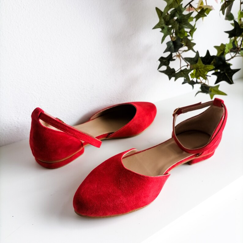 May include: A pair of red suede ballet flats with ankle straps. The flats have a pointed toe and a low heel.