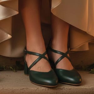 May include: Dark green leather shoes with crisscross straps and a small block heel. The shoes have a closed toe and an ankle strap with a buckle. The shoes are on a stone surface.