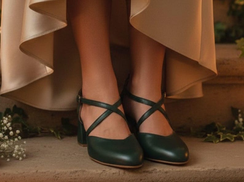 May include: Dark green leather shoes with crisscross straps and a small block heel. The shoes have a closed toe and an ankle strap with a buckle. The shoes are on a stone surface.