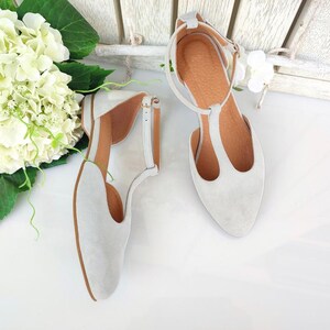 May include: A pair of light gray suede T-strap flats with a pointed toe. The shoes have a small heel and a buckle closure. The interior is a light brown color. The shoes are displayed on a white surface.