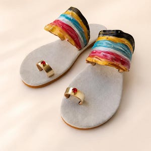 May include: A pair of flat sandals featuring light gray suede insoles and a gold-toned toe ring accented with a single red gemstone. The wide upper strap displays a vibrant, wavy pattern of black, yellow, light blue, and magenta stripes.