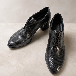 May include: A pair of black leather oxford shoes with a snakeskin pattern. The shoes have a lace-up closure and a small block heel. The shoes are displayed on a neutral background.