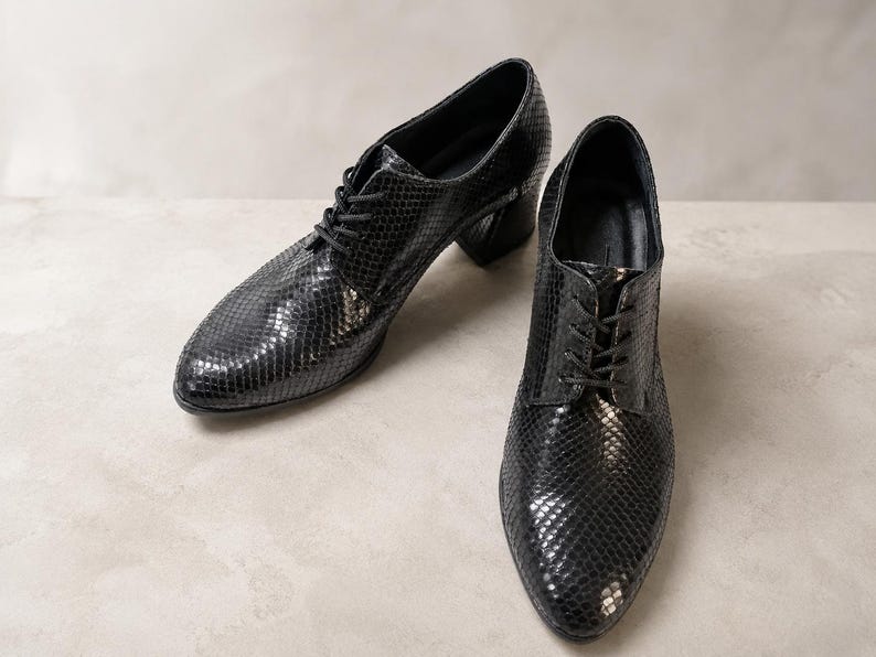 May include: A pair of black leather oxford shoes with a snakeskin pattern. The shoes have a lace-up closure and a small block heel. The shoes are displayed on a neutral background.