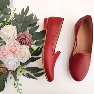 Puede incluir: Un par de mocasines de cuero rojo con tac&oacute;n bajo, exhibidos junto a un arreglo floral de rosas blancas, rosas y malvas con follaje verde. Los zapatos tienen un dise&ntilde;o cl&aacute;sico sin cordones.