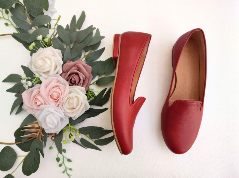 Puede incluir: Un par de mocasines de cuero rojo con tac&oacute;n bajo, exhibidos junto a un arreglo floral de rosas blancas, rosas y malvas con follaje verde. Los zapatos tienen un dise&ntilde;o cl&aacute;sico sin cordones.