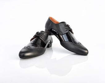 May include: A pair of black dress shoes featuring a combination of matte and patent leather finishes. These lace-up shoes have a wingtip toe cap with decorative brogue detailing, a low heel, and a tan interior lining, presented on a white surface.
