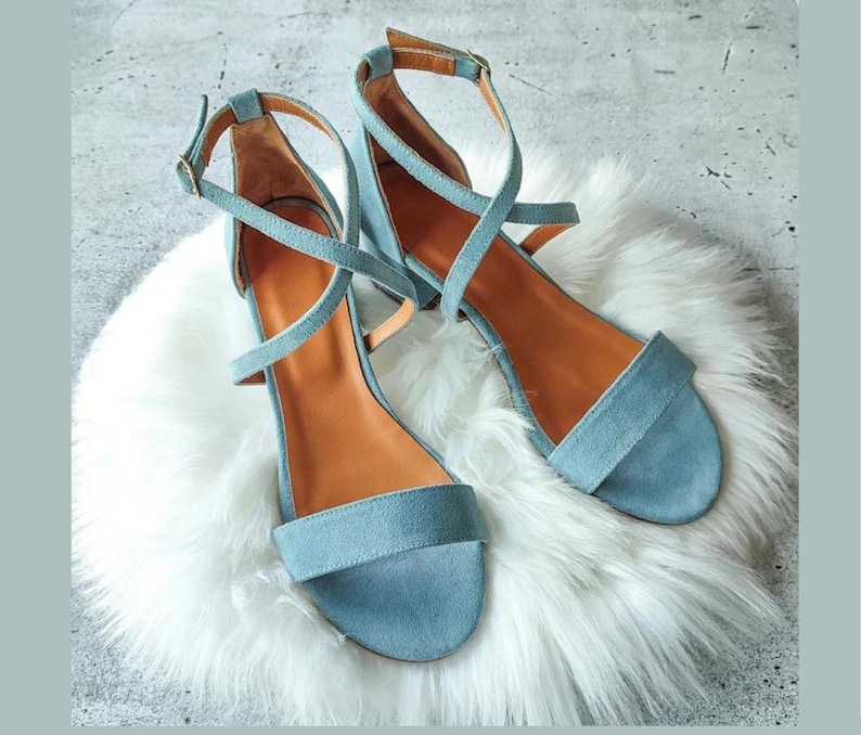 May include: A pair of light blue suede heeled sandals with criss-cross straps. The sandals are on a white fluffy surface.