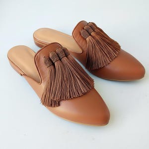 May include: A pair of brown leather mules with a pointed toe and tassel detail. The slip-on shoes have a light brown insole. The tassels are a darker shade of brown. The shoes are set against a white background.