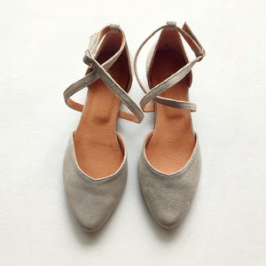 May include: A pair of gray suede ballet flats with criss-cross straps. The straps are tied at the ankle.