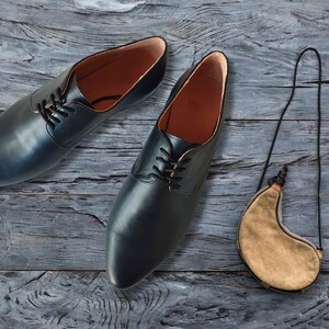 May include: A pair of black leather dress shoes with laces, alongside a tan leather water skin with a black strap. The shoes have a classic design with a rounded toe and a leather interior. The items are arranged on a weathered, gray wooden surface.