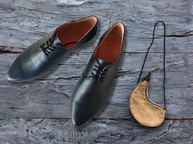 May include: A pair of black leather dress shoes with laces, alongside a tan leather water skin with a black strap. The shoes have a classic design with a rounded toe and a leather interior. The items are arranged on a weathered, gray wooden surface.