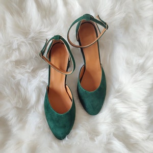 K&ouml;nnte beinhalten: Ein Paar dunkelgr&uuml;ne Wildlederschuhe mit Kn&ouml;chelriemen. Die Schuhe haben eine spitze Zehenpartie und einen geschlossenen R&uuml;cken.