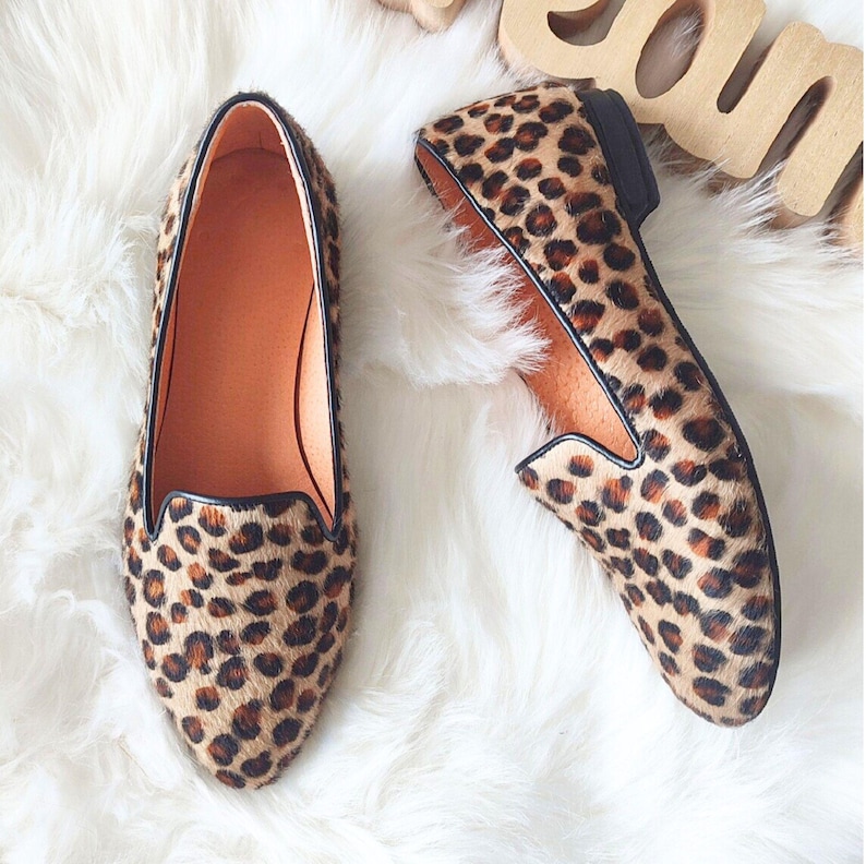 May include: A pair of brown and black leopard print loafers with a black trim around the opening. The loafers are on a white fluffy surface.