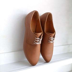 May include: A pair of brown leather oxford shoes with white laces. The shoes have a classic design with a rounded toe and a low heel. The shoes are displayed against a white background.