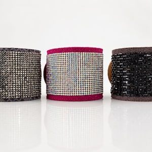 May include: Three wide cuff bracelets arranged in a row. The left cuff is grey with dark metallic studs. The center cuff is bright pink with iridescent rhinestones. The right cuff is brown with a grid of black rectangular beads and studs.