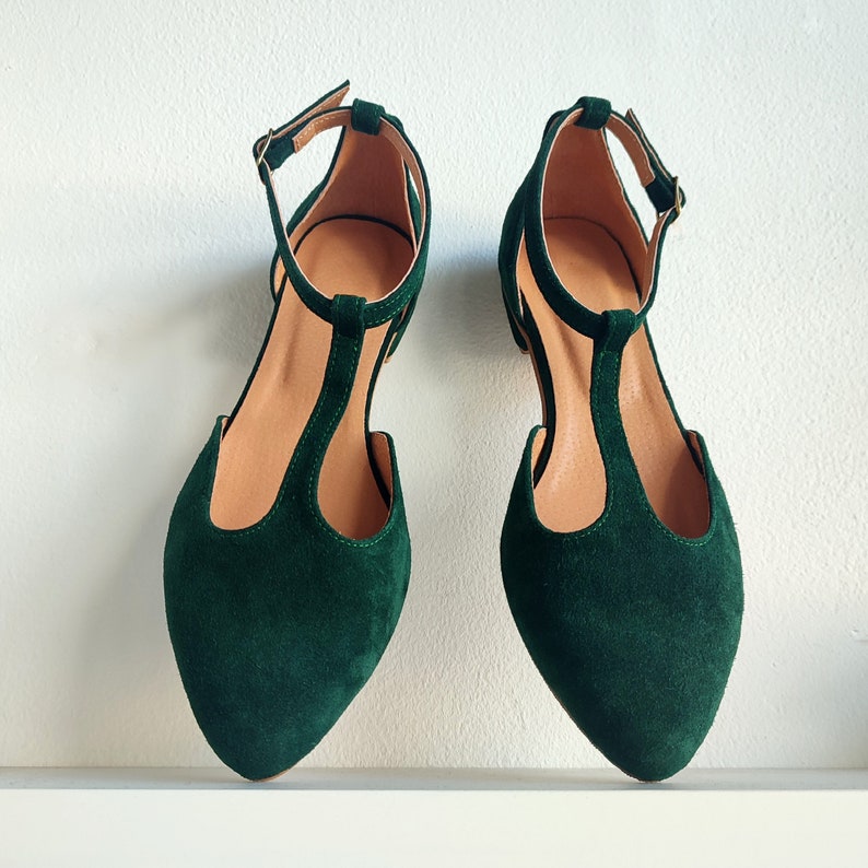 May include: A pair of dark green suede T-strap flats with pointed toes. The shoes have a thin ankle strap with a gold buckle and a light brown insole. The shoes are displayed on a white surface against a white background.