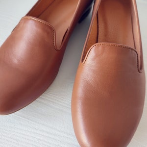 Puede incluir: Primer plano de dos mocasines de cuero marr&oacute;n. Los zapatos tienen un dise&ntilde;o cl&aacute;sico con punta redondeada y tac&oacute;n bajo. El cuero tiene una textura suave y un color marr&oacute;n rojizo c&aacute;lido. Las costuras son visibles a lo largo de los bordes de los zapatos.