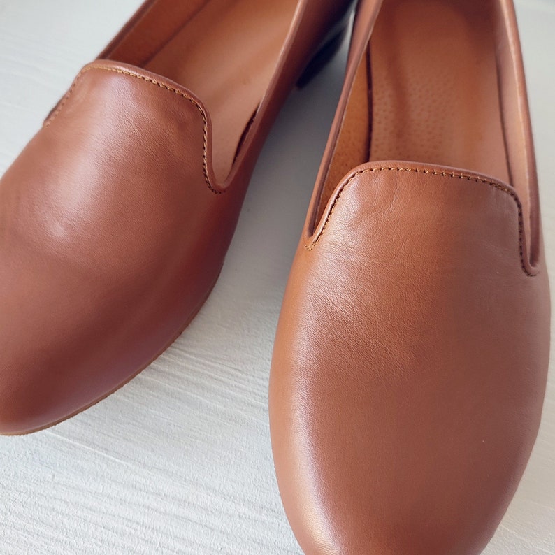 Puede incluir: Primer plano de dos mocasines de cuero marr&oacute;n. Los zapatos tienen un dise&ntilde;o cl&aacute;sico con punta redondeada y tac&oacute;n bajo. El cuero tiene una textura suave y un color marr&oacute;n rojizo c&aacute;lido. Las costuras son visibles a lo largo de los bordes de los zapatos.