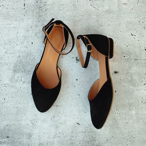 May include: A pair of black suede ankle strap flats with gold buckles. The shoes are on a light gray background.