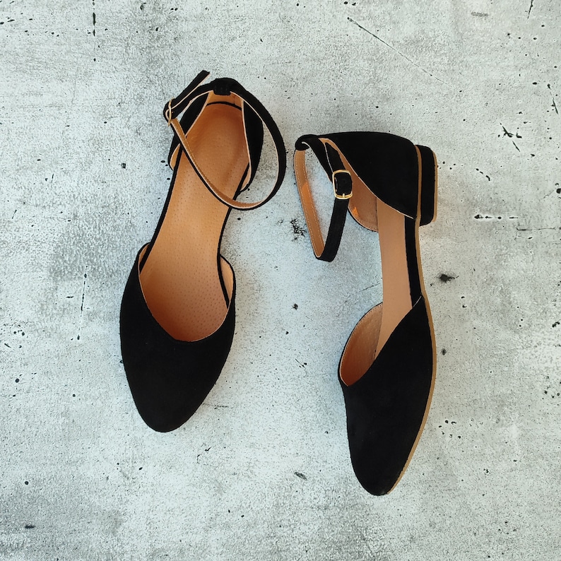 May include: A pair of black suede ankle strap flats with gold buckles. The shoes are on a light gray background.