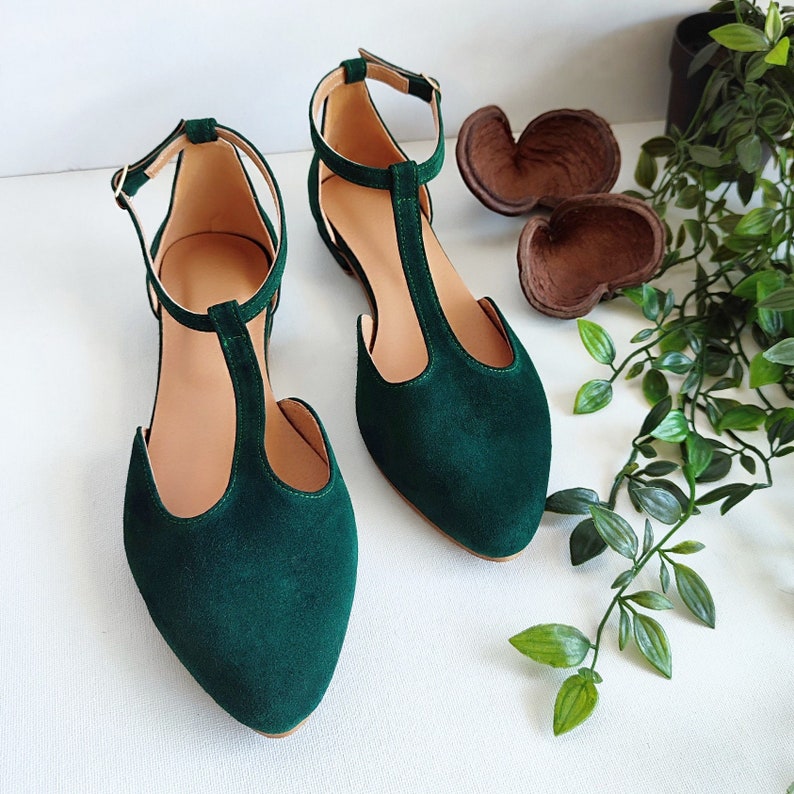 May include: A pair of emerald green T-strap flats with pointed toes. The shoes have a delicate ankle strap with a small gold buckle. The interior of the shoe is a light tan color. The shoes are displayed on a white surface.