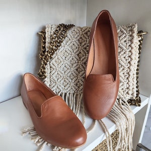 Puede incluir: Dos mocasines de cuero marr&oacute;n se exhiben en una estanter&iacute;a blanca. Los zapatos tienen un dise&ntilde;o cl&aacute;sico con punta redondeada y tac&oacute;n bajo. Una bolsa de macram&eacute; beige con una correa de cadena dorada est&aacute; en el fondo.