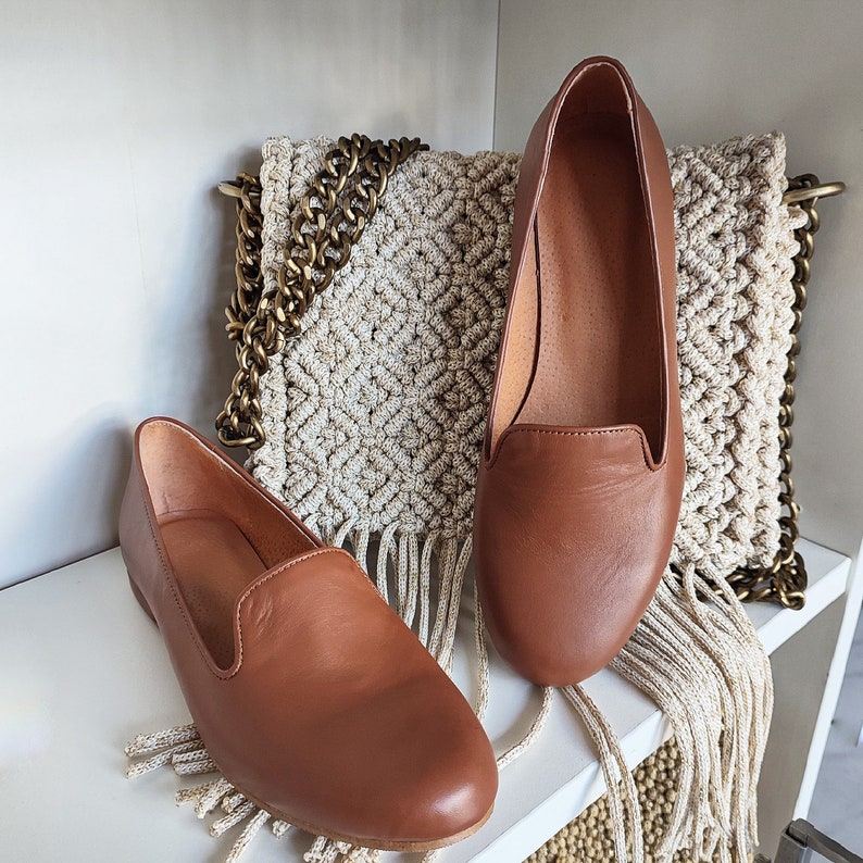 Puede incluir: Dos mocasines de cuero marr&oacute;n se exhiben en una estanter&iacute;a blanca. Los zapatos tienen un dise&ntilde;o cl&aacute;sico con punta redondeada y tac&oacute;n bajo. Una bolsa de macram&eacute; beige con una correa de cadena dorada est&aacute; en el fondo.