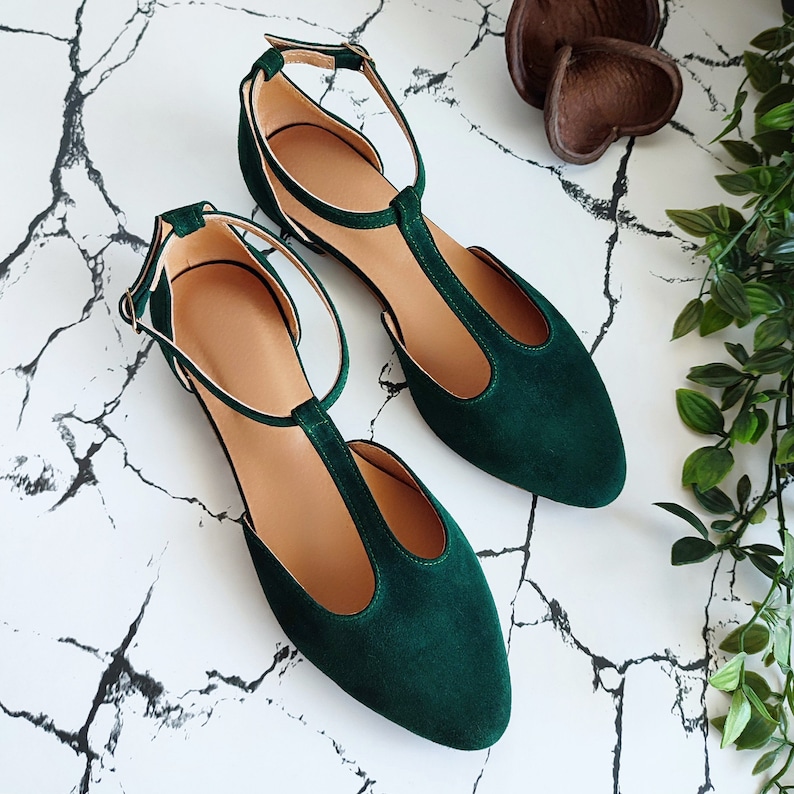 May include: A pair of emerald green T-strap flats with ankle straps. The shoes have a pointed toe and a suede-like texture. The background is a white marble pattern with some greenery and decorative elements.