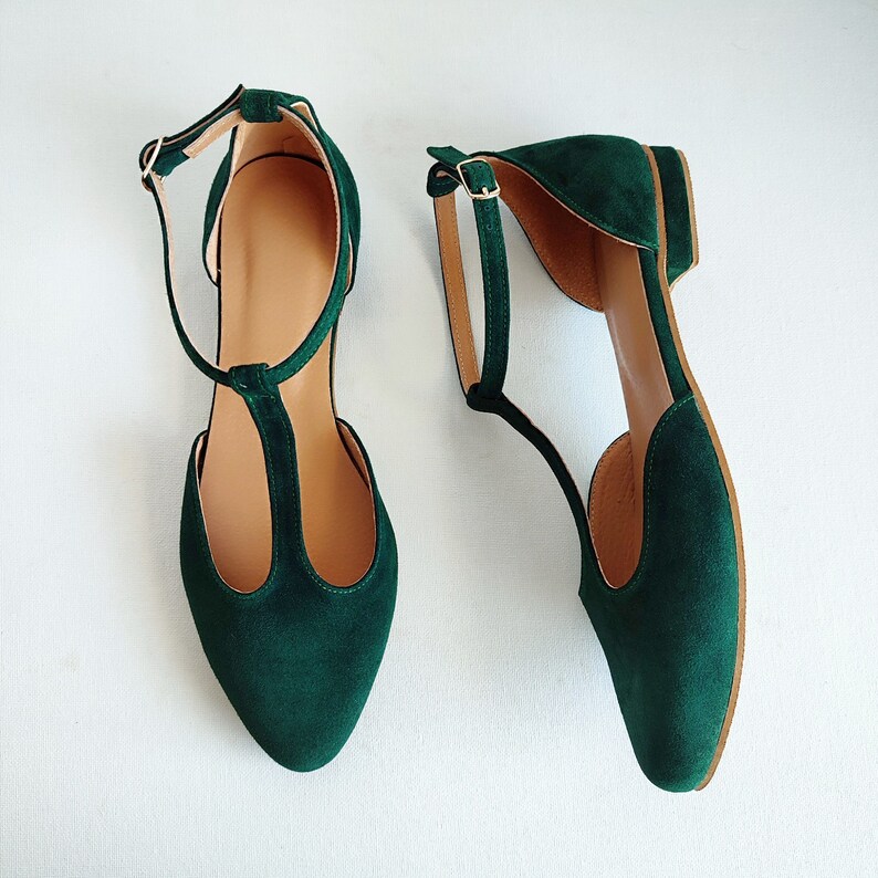May include: A pair of emerald green suede flats with a T-strap design and ankle straps. The shoes have a pointed toe and a small heel. The interior is a light tan color, and the shoes are set against a white background.