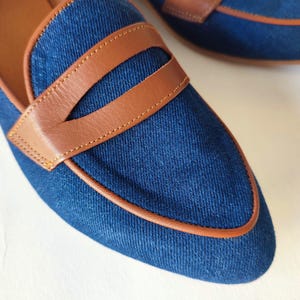May include: A pair of blue denim loafers with brown leather trim. The loafers have a pointed toe and a leather sole.