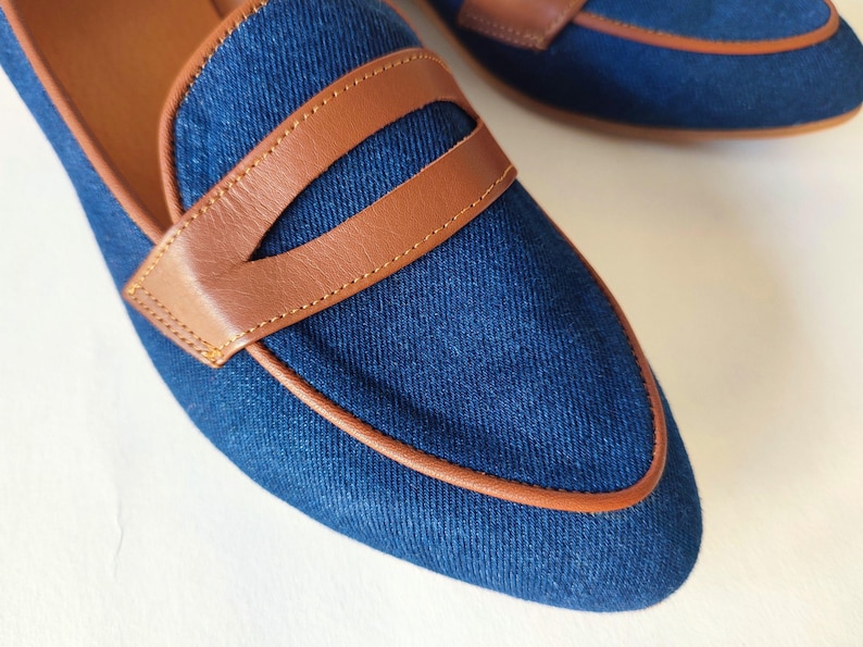 May include: A pair of blue denim loafers with brown leather trim. The loafers have a pointed toe and a leather sole.