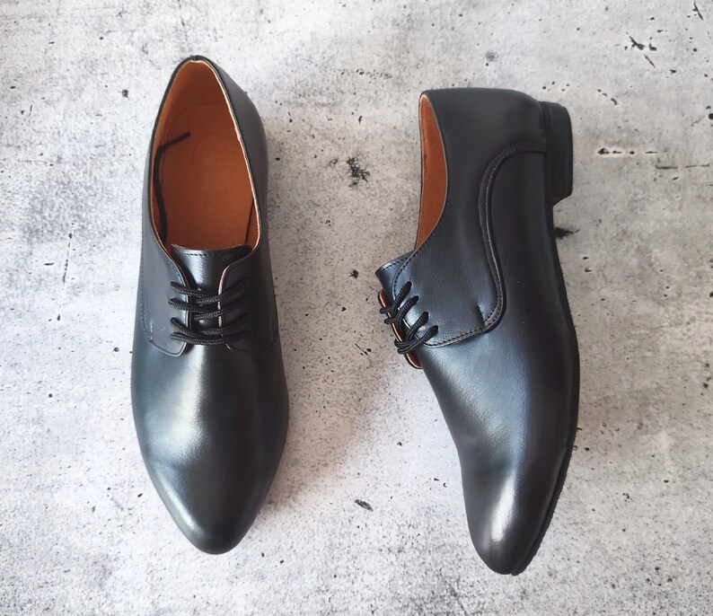 May include: A pair of black leather dress shoes with laces, displayed on a gray surface. The shoes have a classic design with a pointed toe and a low heel. The interior of the shoes is a light brown color.