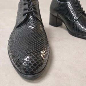 May include: Black leather oxford shoes with a textured snakeskin pattern. The shoes have a lace-up closure and a block heel. The shoes are displayed on a neutral background.