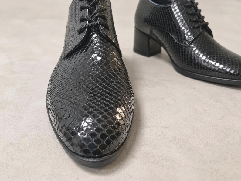 May include: Black leather oxford shoes with a textured snakeskin pattern. The shoes have a lace-up closure and a block heel. The shoes are displayed on a neutral background.