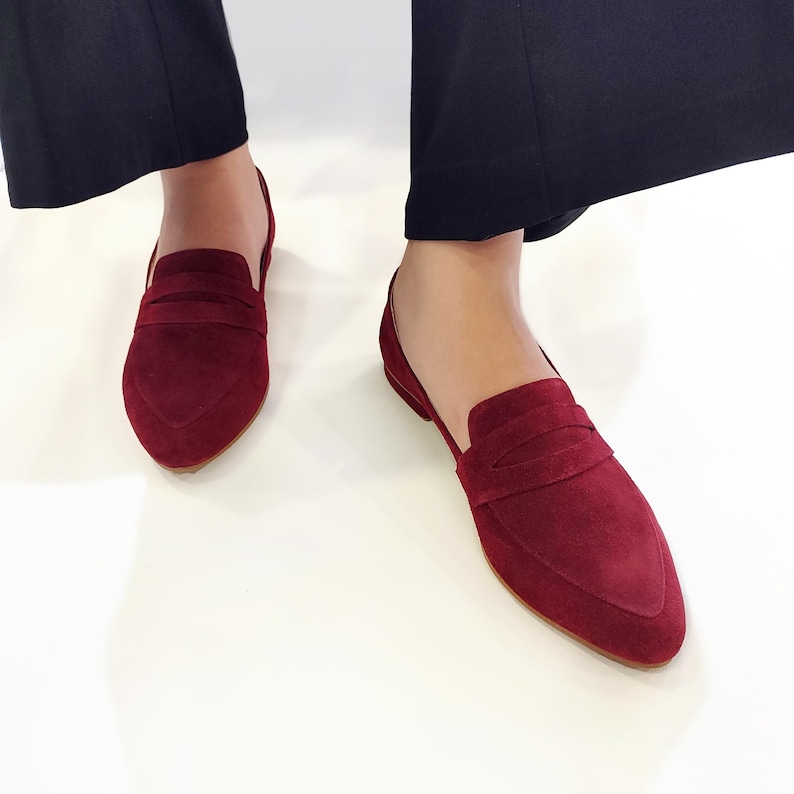 May include: A pair of burgundy suede loafers with a pointed toe and a single strap across the vamp.