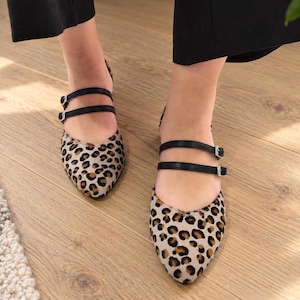 May include: A pair of pointed-toe flats featuring a beige and brown leopard print pattern. Each shoe has two thin black straps across the top, secured with small silver-toned metal buckles, creating a double-strap design over the foot.