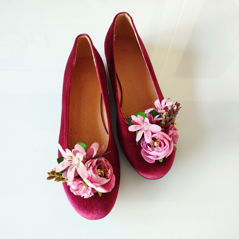 May include: A pair of burgundy velvet flats with pink flower embellishments. The flowers are arranged in a cluster on the toe of each shoe.