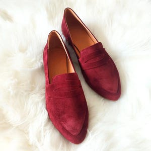 May include: A pair of burgundy suede loafers with a pointed toe and a single strap across the vamp.