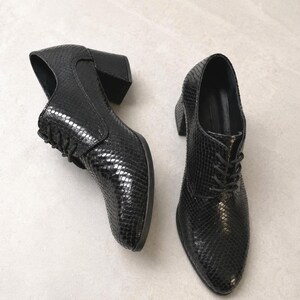 May include: A pair of black leather heeled oxfords with a textured snakeskin pattern. The shoes have a lace-up design and a block heel. The shoes are displayed on a light gray surface.