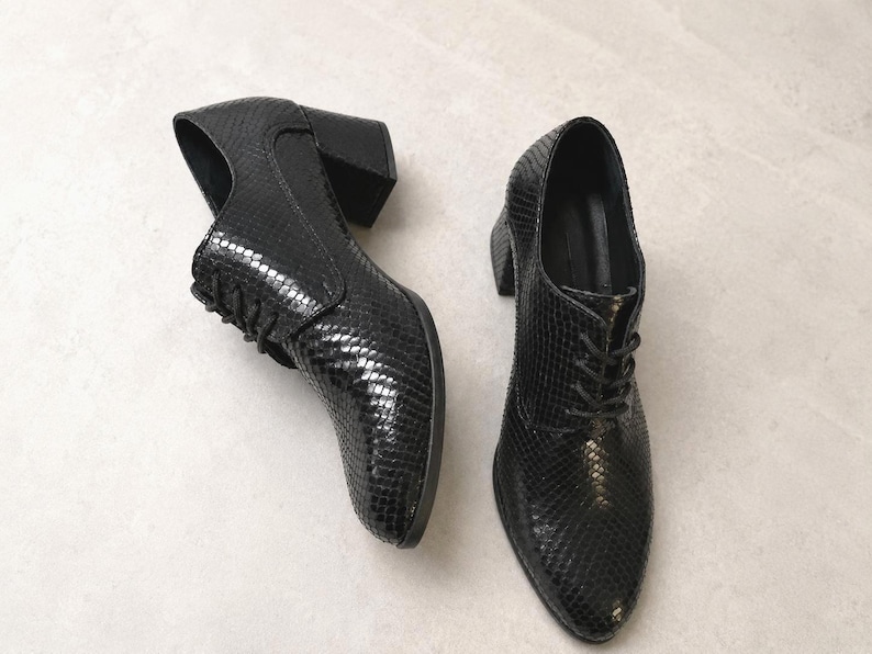 May include: A pair of black leather heeled oxfords with a textured snakeskin pattern. The shoes have a lace-up design and a block heel. The shoes are displayed on a light gray surface.