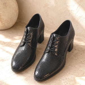 May include: A pair of black, heeled oxford shoes with a textured, snakeskin-like pattern. The shoes have a lace-up closure and a chunky heel. The shoes are displayed on a light-colored surface.