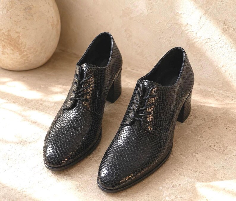 May include: A pair of black, heeled oxford shoes with a textured, snakeskin-like pattern. The shoes have a lace-up closure and a chunky heel. The shoes are displayed on a light-colored surface.