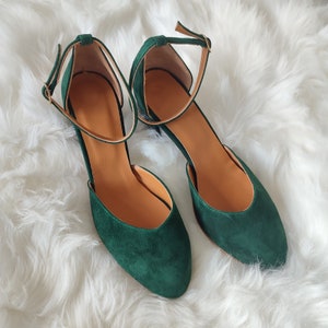 K&ouml;nnte beinhalten: Ein Paar dunkelgr&uuml;ne Wildlederschuhe mit Kn&ouml;chelriemen. Die Schuhe haben eine spitze Zehenpartie und einen niedrigen Blockabsatz.