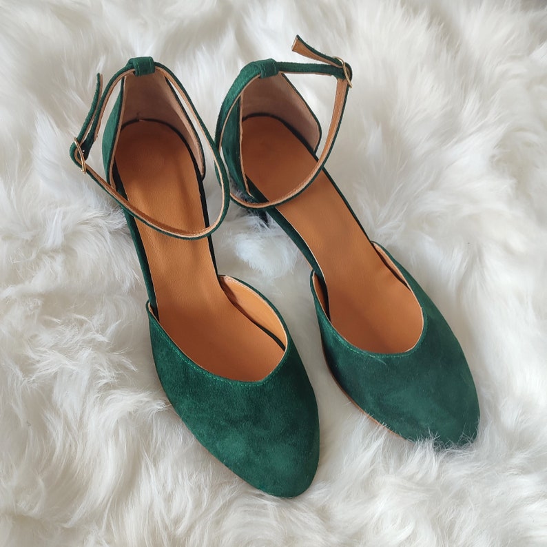 K&ouml;nnte beinhalten: Ein Paar dunkelgr&uuml;ne Wildlederschuhe mit Kn&ouml;chelriemen. Die Schuhe haben eine spitze Zehenpartie und einen niedrigen Blockabsatz.