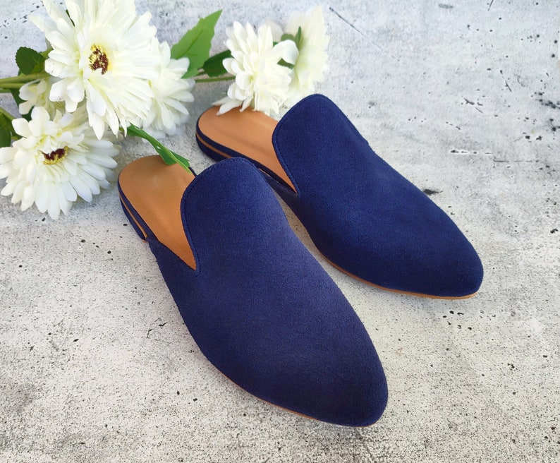 May include: A pair of navy blue suede slip-on shoes with a tan leather interior. The shoes are displayed on a light gray surface, with white flowers in the background. The shoes have a classic, minimalist design.