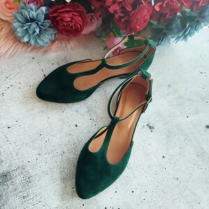 May include: A pair of emerald green suede flats with a T-strap design. The shoes have a pointed toe and a delicate ankle strap with a gold-colored buckle. The interior of the shoe is a light tan color.