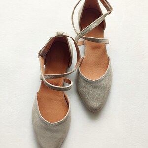 May include: A pair of gray suede ballet flats with ankle straps. The straps crisscross over the top of the foot and are secured with a buckle.