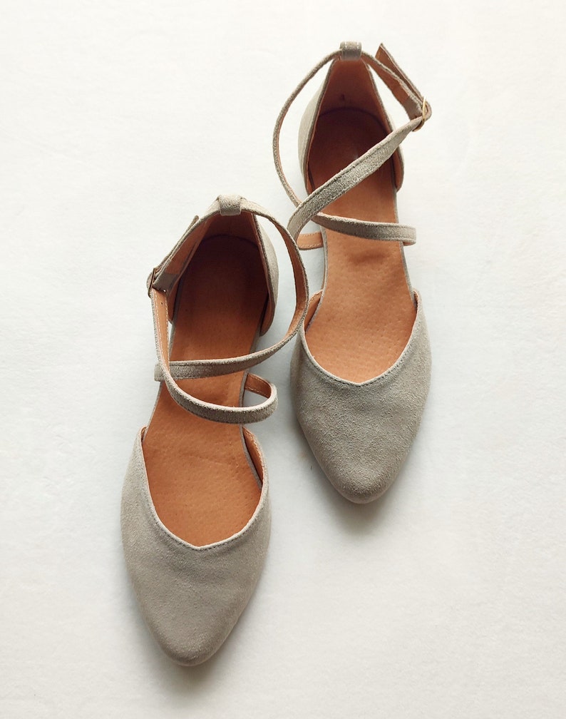 May include: A pair of gray suede ballet flats with ankle straps. The straps crisscross over the top of the foot and are secured with a buckle.
