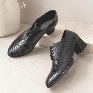 May include: A pair of black, textured leather shoes with a lace-up design and block heels. The shoes have a snakeskin pattern and are displayed on a neutral surface. The heels appear to be approximately 2 inches high.